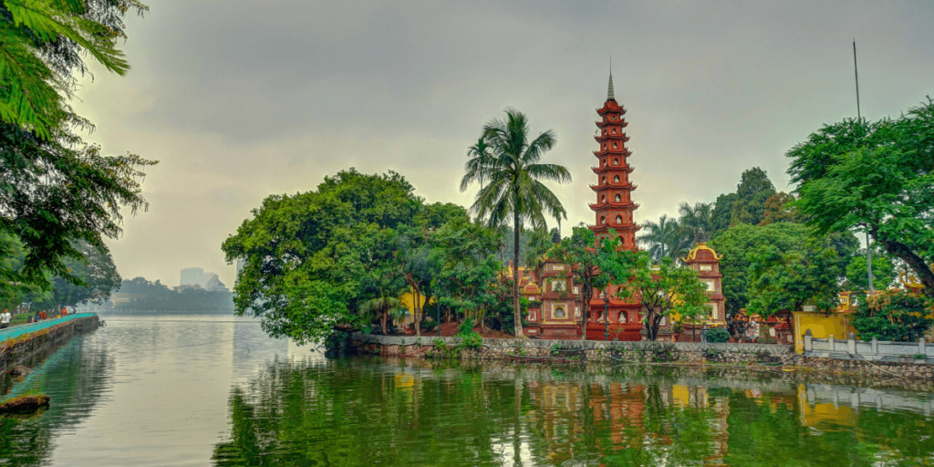 這張圖片展示了越南河內的獨柱寺（One Pillar Pagoda）。這座寺廟建築獨特，建在單一的柱子上，四周是綠樹和池塘。寺廟的背景被寧靜的自然景觀包圍，展示了越南古老的文化與宗教特色。