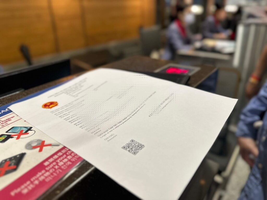 這張圖片顯示了一張越南電子簽證文件,放置在登機櫃台的桌面上。文件的上方印有越南的國徽,顯示出這是正式的入境許可證。背景中可以看到正在辦理登機手續的旅客,標誌著這份電子簽證正被用於即將登機的行程。此圖片展示了越南電子簽證在旅行過程中的實際應用,便捷地在登機時進行檢查。